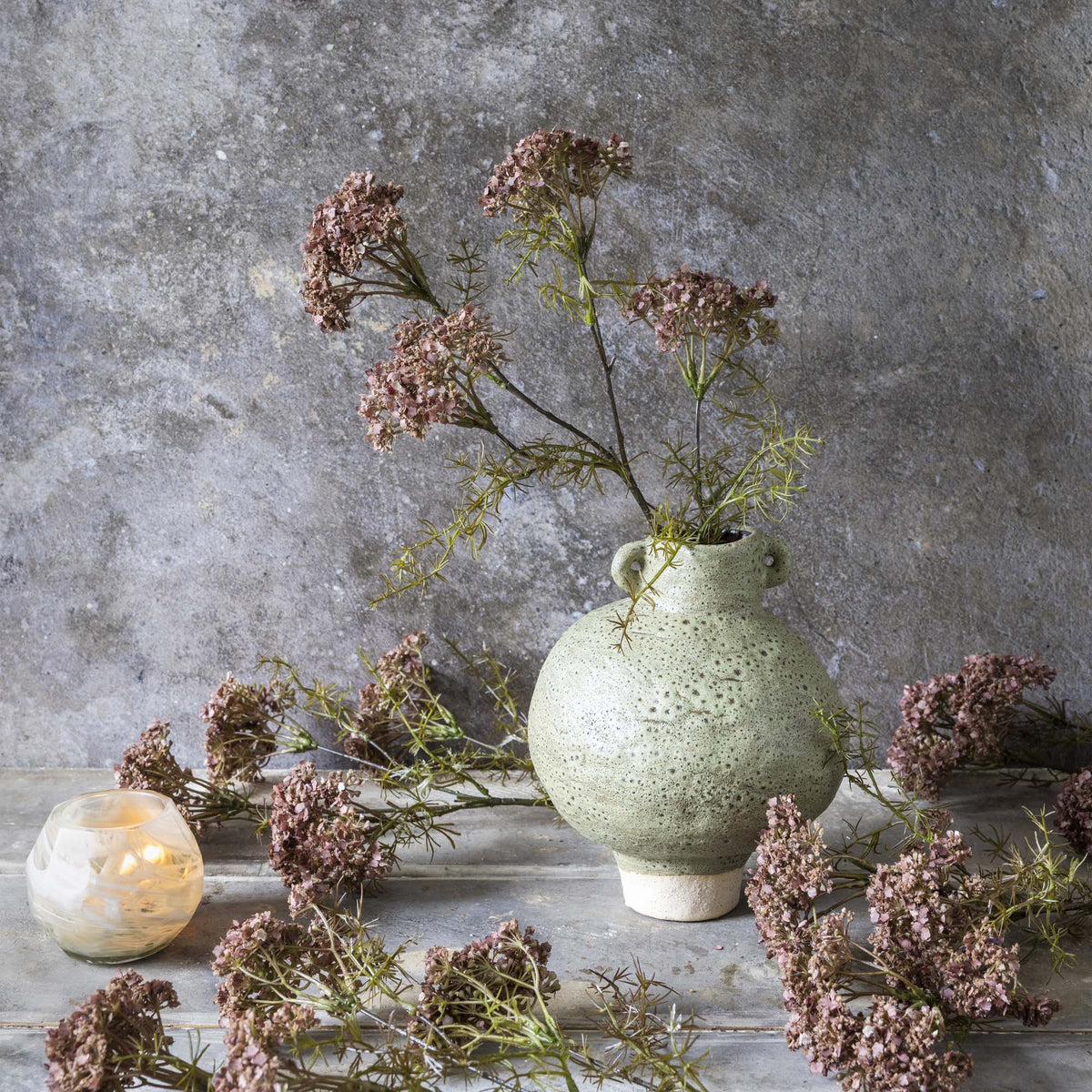 Green ceramic vase with faux flowers on a textured surface against a gray wall.